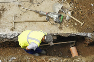 Plumber with Digging Tools in Trench Repairing Main Sewer Line